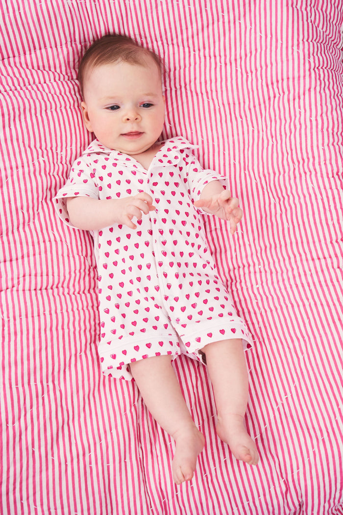 A baby with light skin and light brown hair lies on a pink and white striped blanket wearing the Roller Rabbit Infant Hearts Polo Onesie, looking slightly to the side.