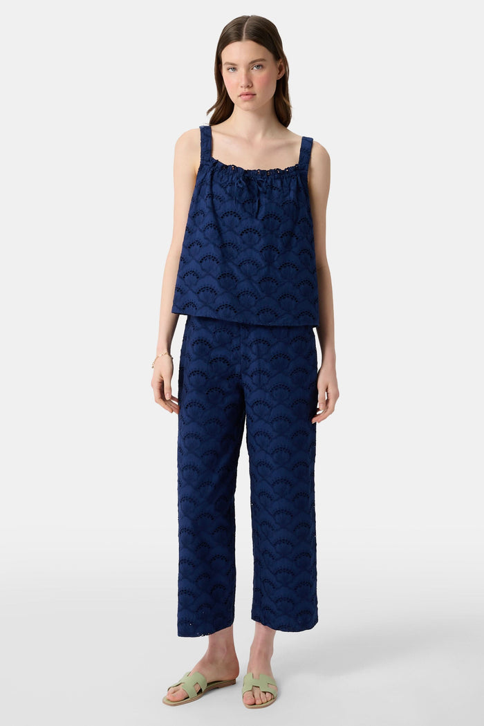 A woman with long brown hair wears the Roller Rabbit Jemina Eyelet Estelle Top in navy and wide-leg pants with a scalloped texture, paired with light green slide sandals. She stands against a plain white background.