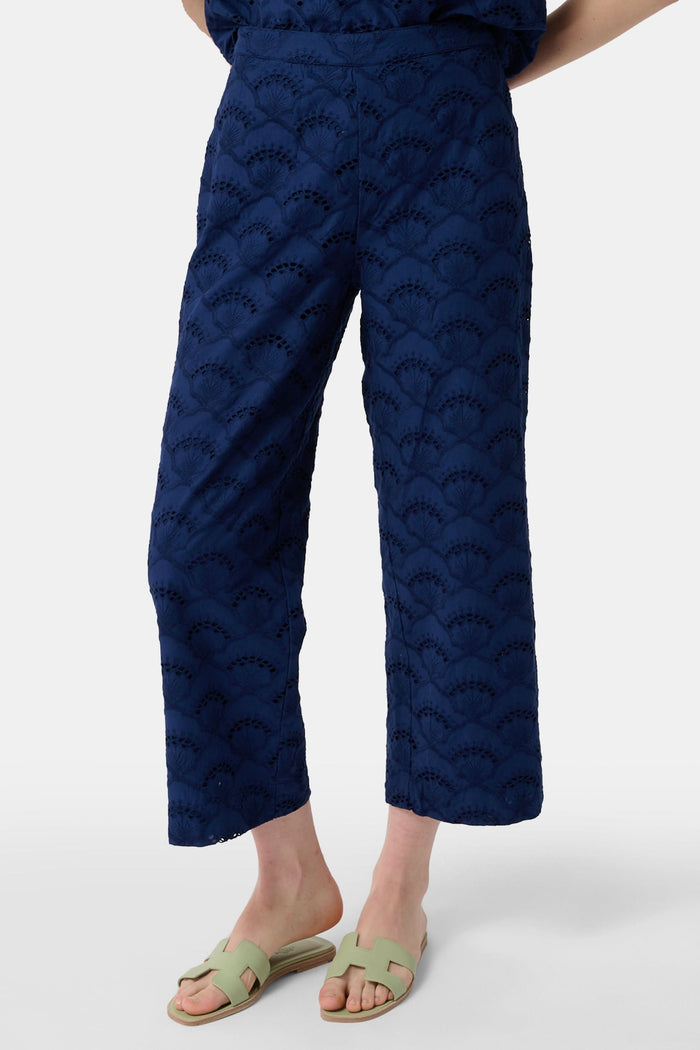 A person wearing Roller Rabbit's Jemina Eyelet Kendall Pant in navy blue, ankle-length, paired with light green slide sandals and standing against a plain white background.