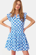 A woman with long, wavy light brown hair stands against a white background, wearing the Roller Rabbit Jemina Pippa Dress—a short-sleeved cotton voile dress in blue Jemina print with a V-neckline and flared skirt. She poses with hands on hips and a neutral expression.