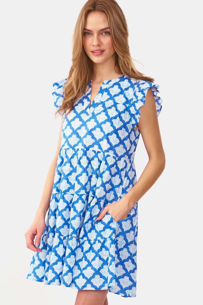 A woman with long, wavy light brown hair stands against a white background, wearing the Roller Rabbit Jemina Pippa Dress—a short-sleeved cotton voile dress in blue Jemina print with a V-neckline and flared skirt. She poses with hands on hips and a neutral expression.
