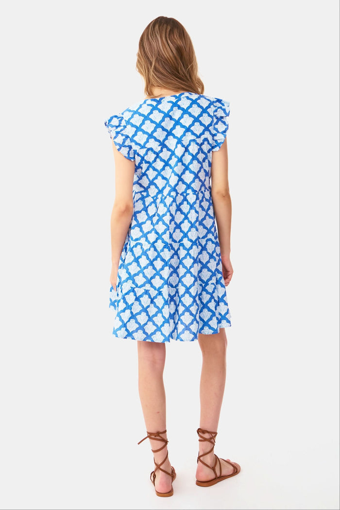 A woman with long, wavy light brown hair stands against a white background, wearing the Roller Rabbit Jemina Pippa Dress—a short-sleeved cotton voile dress in blue Jemina print with a V-neckline and flared skirt. She poses with hands on hips and a neutral expression.