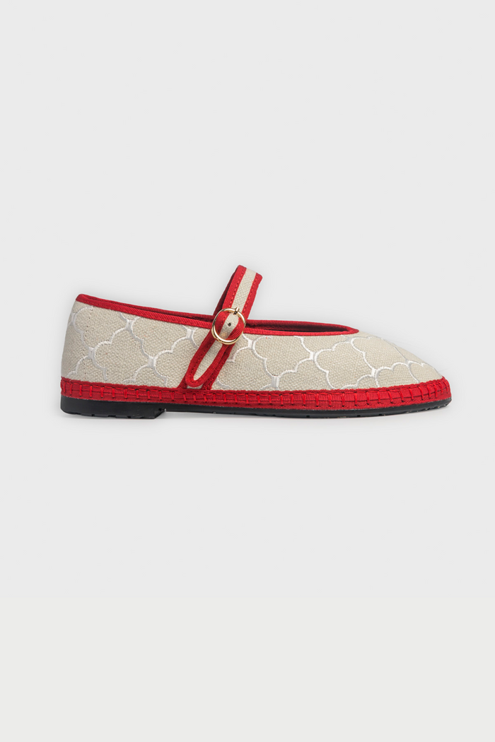 The Jena Embroidery Pierre by FLABELUS is a beige Mary Jane shoe, handmade in Spain, with a white pattern, red trim, a red strap and gold buckle, plus a black sole.