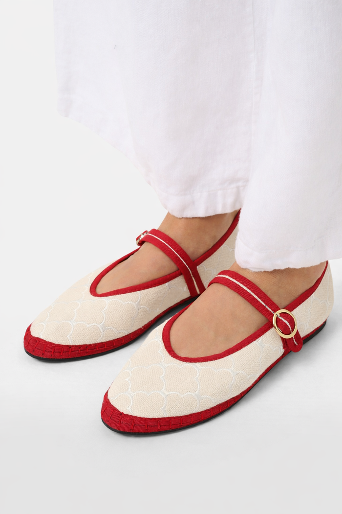 The Jena Embroidery Pierre by FLABELUS is a beige Mary Jane shoe, handmade in Spain, with a white pattern, red trim, a red strap and gold buckle, plus a black sole.