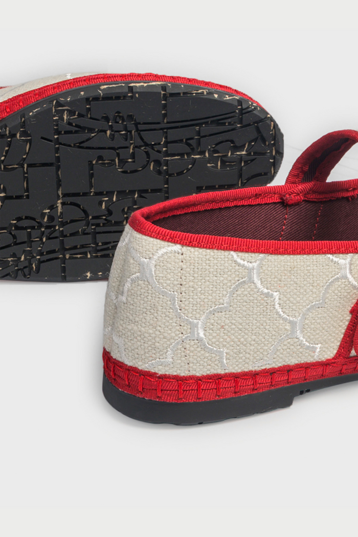 The Jena Embroidery Pierre by FLABELUS is a beige Mary Jane shoe, handmade in Spain, with a white pattern, red trim, a red strap and gold buckle, plus a black sole.