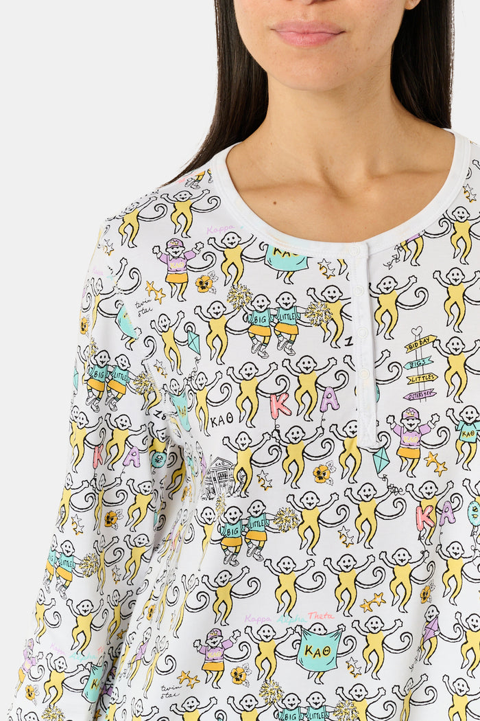 The Roller Rabbit Kappa Alpha Theta Monkey Pajamas set features a white, long-sleeved buttoned top and matching pants, both adorned with a vibrant all-over cartoon print of playful animal characters, including cheerful monkeys.