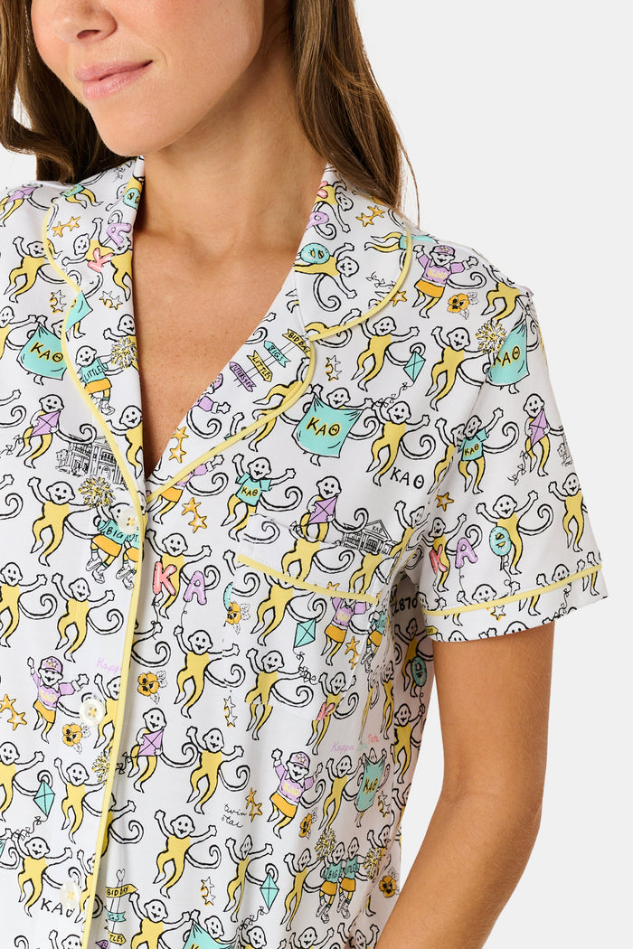 The Kappa Alpha Theta Monkey Polo Pajamas from Roller Rabbit are a short-sleeve button-up set with matching shorts, showcasing a lively print of colorful cartoon monkeys and doodles on a crisp white background. They feature an elastic waistband for comfort and are crafted from soft Pima cotton for a gentle feel.