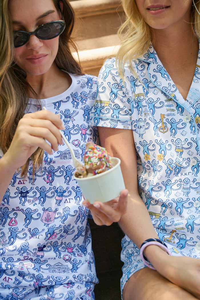 The Roller Rabbit Kappa Kappa Gamma Monkey Polo Pajamas feature a short-sleeve button-up pajama shirt with a front button placket and matching shorts with an elastic waistband, adorned with a playful blue monkey and yellow motif on a white background. Crafted from soft Pima cotton for extra comfort.