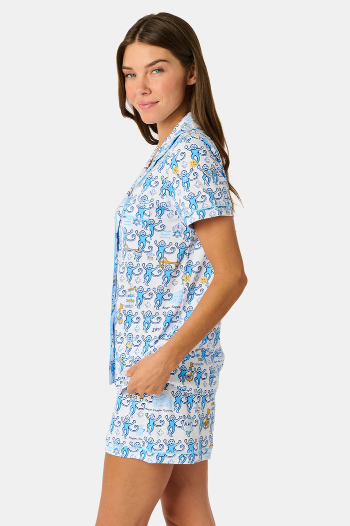 The Roller Rabbit Kappa Kappa Gamma Monkey Polo Pajamas feature a short-sleeve button-up pajama shirt with a front button placket and matching shorts with an elastic waistband, adorned with a playful blue monkey and yellow motif on a white background. Crafted from soft Pima cotton for extra comfort.