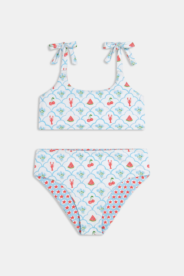 A young girl with curly hair wears the Roller Rabbit Kids 4th Of Jemina/Starry Dot Cora Bikini. She stands against a plain light gray background, looking at the camera with a slight smile.