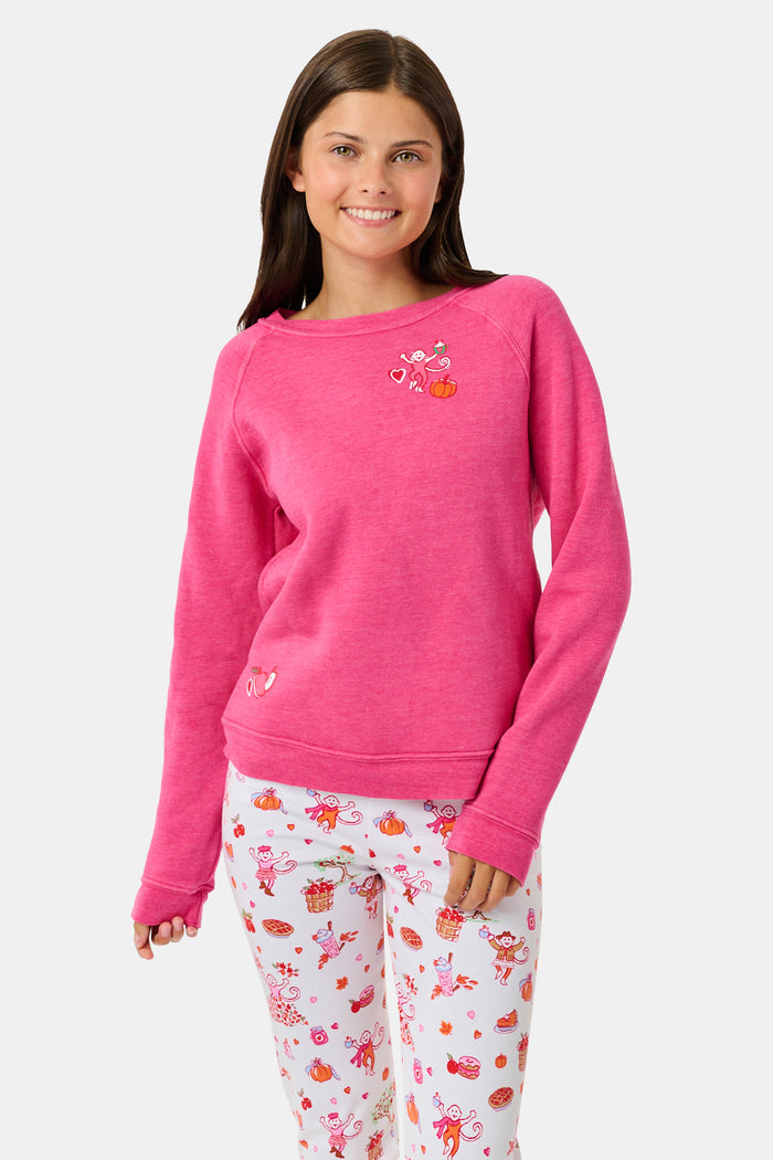 A young girl smiles, wearing the Roller Rabbit Kids Autumn Hearts Crewneck—a pink sweatshirt featuring small embroidered butterflies—paired with a white skirt adorned with a colorful heart pattern, as she stands with her hands behind her back.