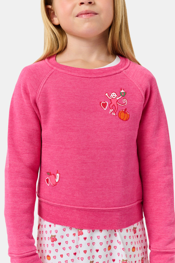 A young girl smiles, wearing the Roller Rabbit Kids Autumn Hearts Crewneck—a pink sweatshirt featuring small embroidered butterflies—paired with a white skirt adorned with a colorful heart pattern, as she stands with her hands behind her back.