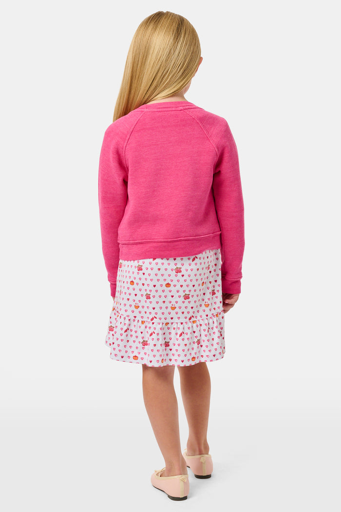 A young girl smiles, wearing the Roller Rabbit Kids Autumn Hearts Crewneck—a pink sweatshirt featuring small embroidered butterflies—paired with a white skirt adorned with a colorful heart pattern, as she stands with her hands behind her back.
