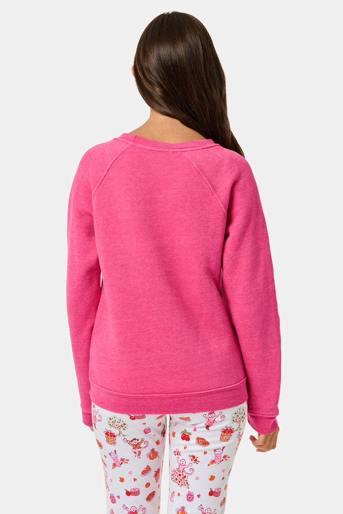 A young girl smiles, wearing the Roller Rabbit Kids Autumn Hearts Crewneck—a pink sweatshirt featuring small embroidered butterflies—paired with a white skirt adorned with a colorful heart pattern, as she stands with her hands behind her back.
