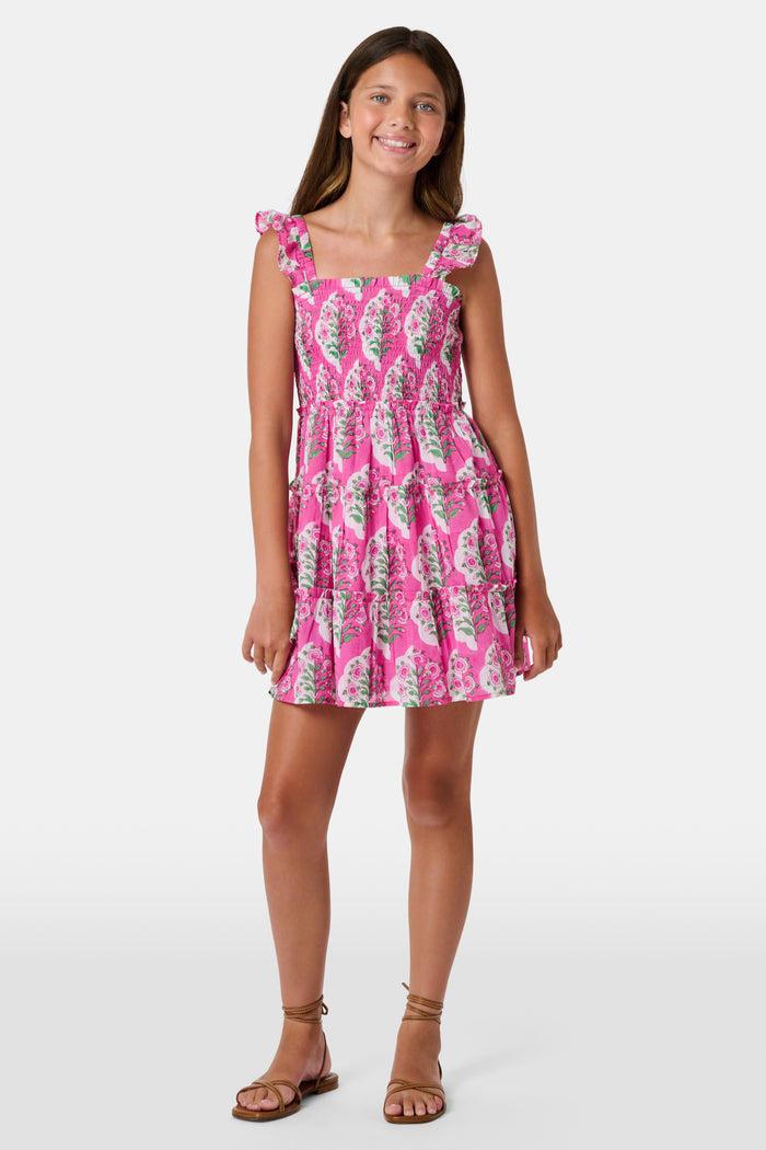 The Kids Ava Floral Ceri Dress by Roller Rabbit is a sleeveless baby dress featuring a smocked bodice and ruffled straps, adorned with green and white leaf patterns on a vibrant pink background, and comes with matching bloomers.
