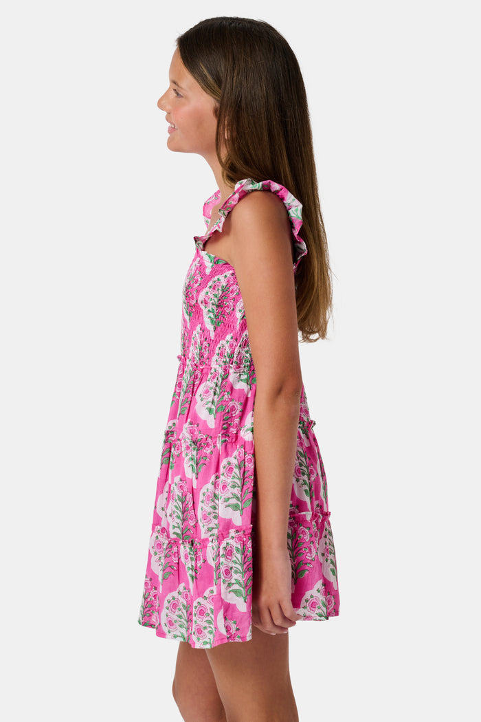 The Kids Ava Floral Ceri Dress by Roller Rabbit is a sleeveless baby dress featuring a smocked bodice and ruffled straps, adorned with green and white leaf patterns on a vibrant pink background, and comes with matching bloomers.