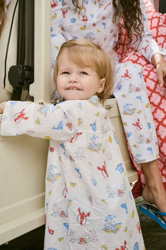 A young child with light brown hair smiles near an open car door, wearing the Roller Rabbit Kids Catch of the Day Serafina Tunic. An adult in matching pajamas is partially visible behind them.