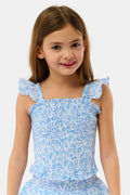 A young girl with long brown hair smiles in the Roller Rabbit Kids Cherry Blossom Toile Tory Top, a light blue and white floral sleeveless cotton voile piece with ruffled shoulder straps, standing against a plain light background.
