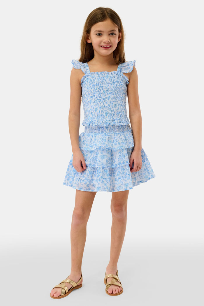 A young girl with long brown hair smiles in the Roller Rabbit Kids Cherry Blossom Toile Tory Top, a light blue and white floral sleeveless cotton voile piece with ruffled shoulder straps, standing against a plain light background.