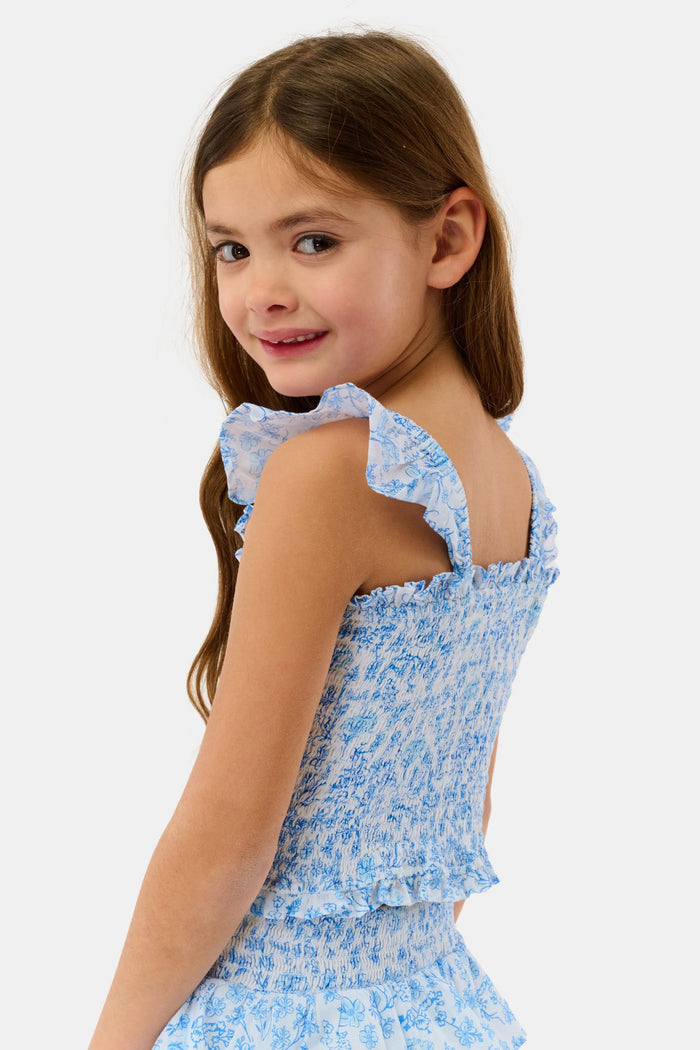A young girl with long brown hair smiles in the Roller Rabbit Kids Cherry Blossom Toile Tory Top, a light blue and white floral sleeveless cotton voile piece with ruffled shoulder straps, standing against a plain light background.