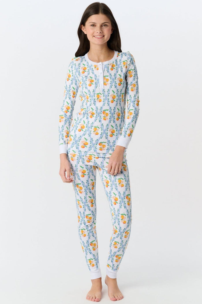 A girl stands barefoot wearing Roller Rabbit Kids Clementine Trellis Pajamas, featuring an elastic waist and a vibrant orange and blue floral fruit pattern on a white background, smiling with her arms at her sides.