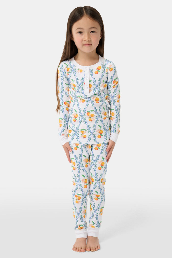 A girl stands barefoot wearing Roller Rabbit Kids Clementine Trellis Pajamas, featuring an elastic waist and a vibrant orange and blue floral fruit pattern on a white background, smiling with her arms at her sides.