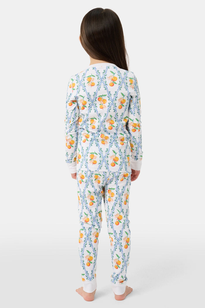 A girl stands barefoot wearing Roller Rabbit Kids Clementine Trellis Pajamas, featuring an elastic waist and a vibrant orange and blue floral fruit pattern on a white background, smiling with her arms at her sides.