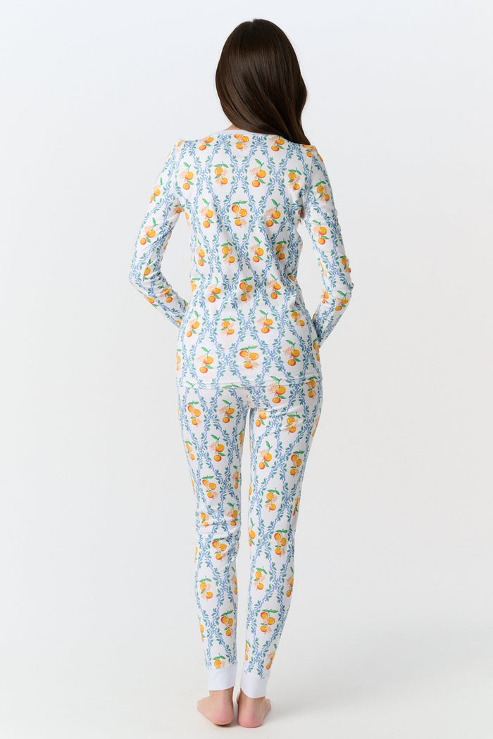A girl stands barefoot wearing Roller Rabbit Kids Clementine Trellis Pajamas, featuring an elastic waist and a vibrant orange and blue floral fruit pattern on a white background, smiling with her arms at her sides.