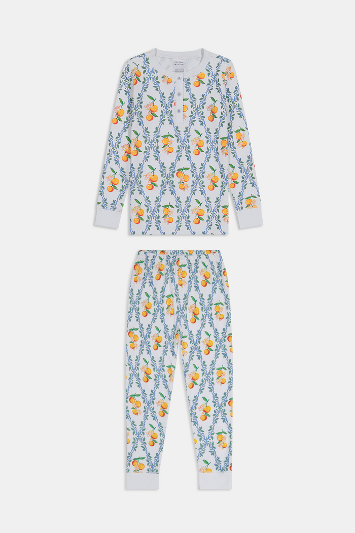 A girl stands barefoot wearing Roller Rabbit Kids Clementine Trellis Pajamas, featuring an elastic waist and a vibrant orange and blue floral fruit pattern on a white background, smiling with her arms at her sides.