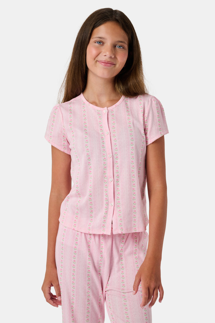 A young girl in a rose pink dress smiles while kneeling on the floor, holding a colorful folder. A pink backpack hangs nearby in a classroom or locker area, and she wears her cozy Roller Rabbit Kids Daisy Heart Stripe Diana Cardigan.
