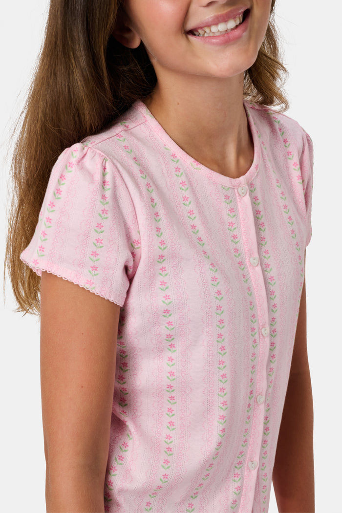 A young girl in a rose pink dress smiles while kneeling on the floor, holding a colorful folder. A pink backpack hangs nearby in a classroom or locker area, and she wears her cozy Roller Rabbit Kids Daisy Heart Stripe Diana Cardigan.
