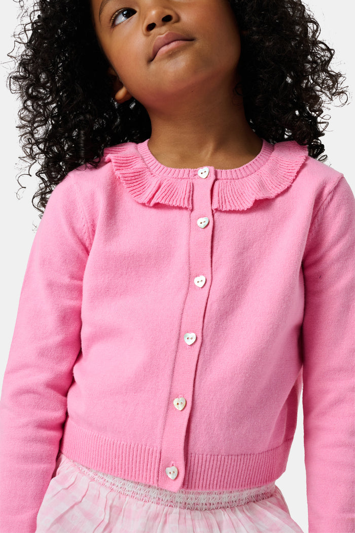 The Kids Ellis Cardigan by Roller Rabbit is a rose pink, long-sleeve cardigan featuring a ruffled collar and pearl-like buttons. Shown flat on a white background, it's made from soft cotton sweater material.