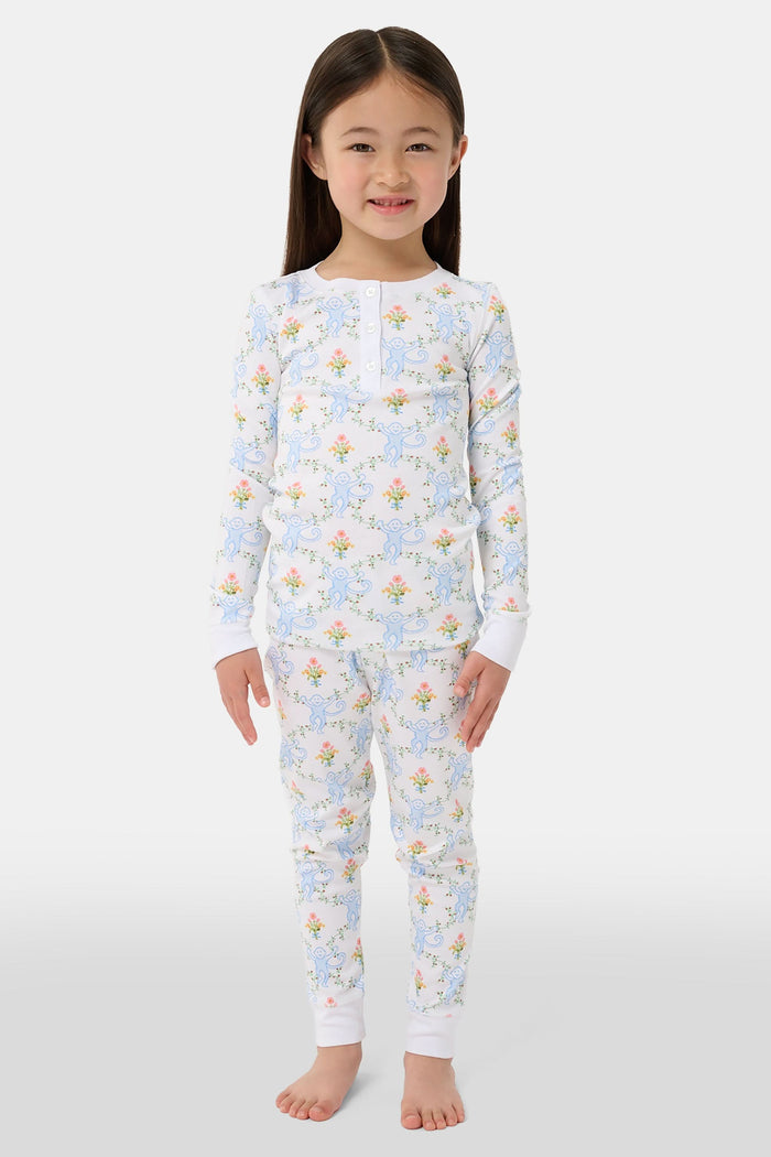 A child in Kids Floral Monkey Trellis Pajamas by Roller Rabbit sits barefoot on an adult’s lap outdoors. The adult, also barefoot and wearing light blue Pima Cotton sleepwear, hugs the child gently; only part of them is visible.