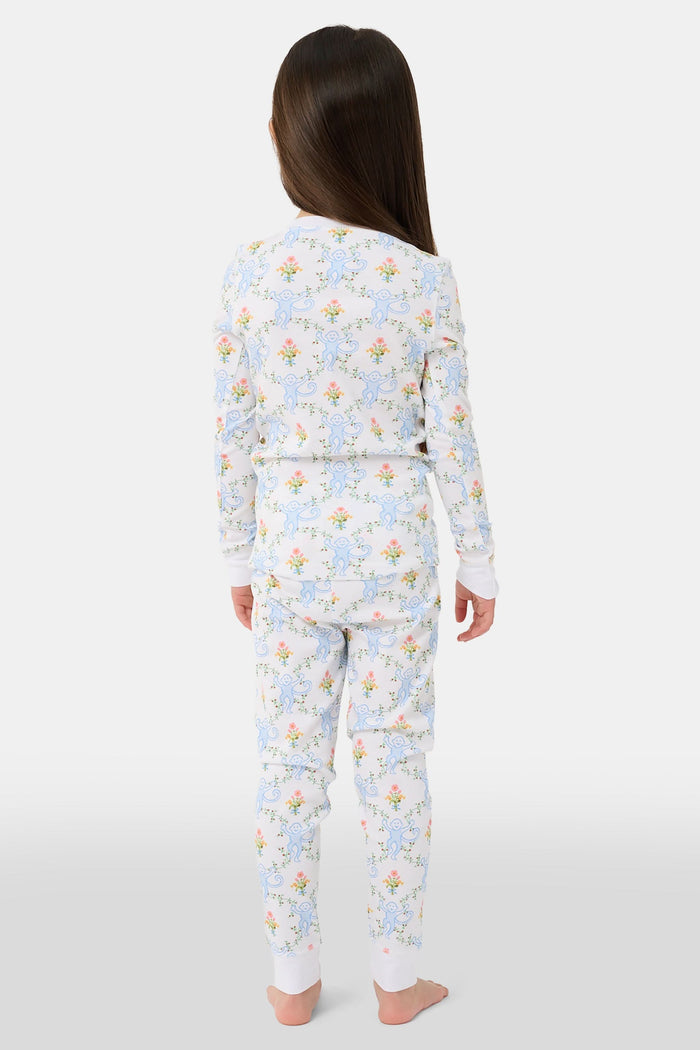 A child in Kids Floral Monkey Trellis Pajamas by Roller Rabbit sits barefoot on an adult’s lap outdoors. The adult, also barefoot and wearing light blue Pima Cotton sleepwear, hugs the child gently; only part of them is visible.
