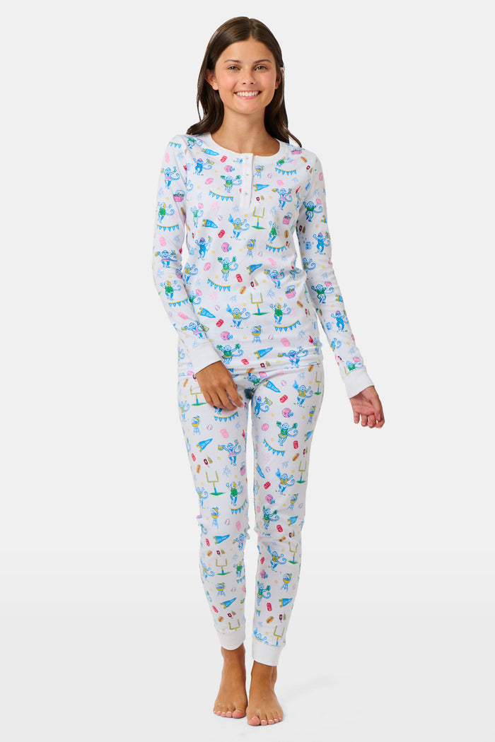 The Kids Game Day Pajamas by Roller Rabbit are a children’s long-sleeve pajama set crafted from soft Pima cotton, featuring a vibrant party-themed print with colorful dinosaurs, bunting, gifts, and party hats on a white background.