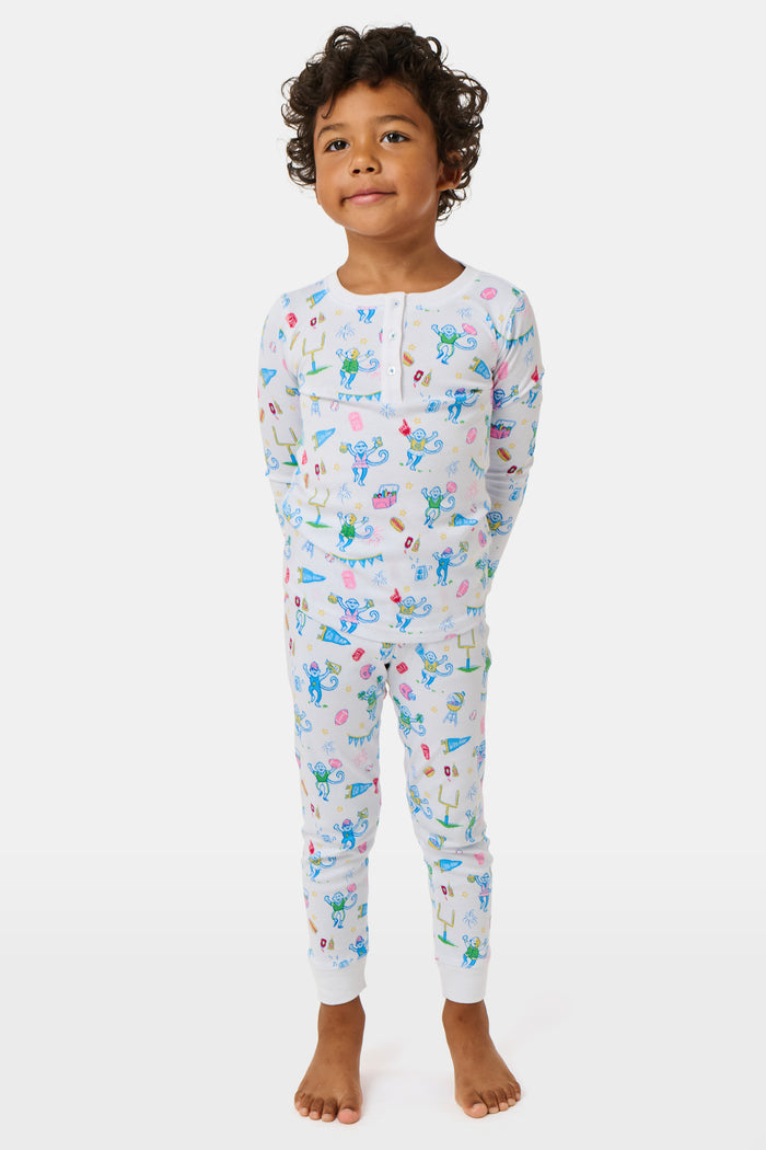 The Kids Game Day Pajamas by Roller Rabbit are a children’s long-sleeve pajama set crafted from soft Pima cotton, featuring a vibrant party-themed print with colorful dinosaurs, bunting, gifts, and party hats on a white background.