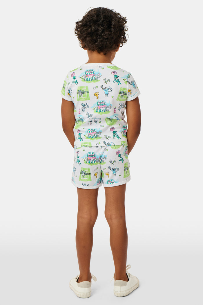 A girl stands outdoors in a Roller Rabbit Kids Game, Set, Monkey Playa Play Set, holding out a tennis ball toward the camera. Her playful outfit is perfect for kids’ tennis activities, and the photo is taken from a low angle with blue sky and trees in the background.