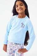 A young girl with long dark hair smiles, posing with one hand on her hip. She wears the Roller Rabbit Kids Georgetown Monkey Crewneck sweatshirt in light blue and colorful patterned leggings with unicorns and rainbows.