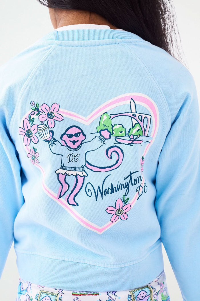 A young girl with long dark hair smiles, posing with one hand on her hip. She wears the Roller Rabbit Kids Georgetown Monkey Crewneck sweatshirt in light blue and colorful patterned leggings with unicorns and rainbows.