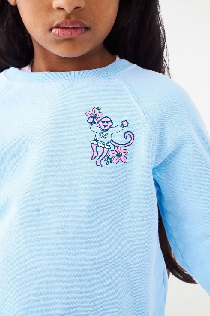 A young girl with long dark hair smiles, posing with one hand on her hip. She wears the Roller Rabbit Kids Georgetown Monkey Crewneck sweatshirt in light blue and colorful patterned leggings with unicorns and rainbows.