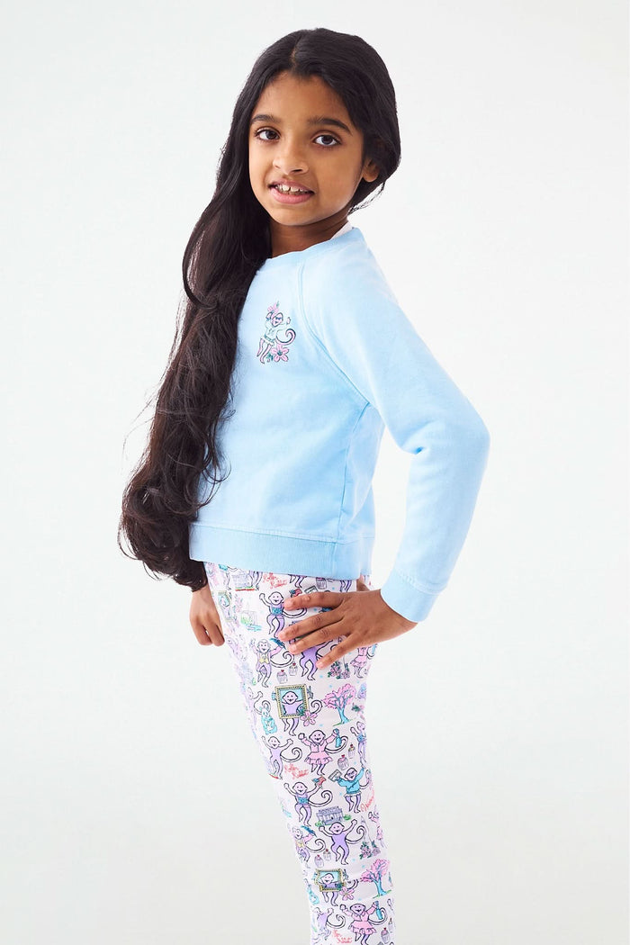 A young girl with long dark hair smiles, posing with one hand on her hip. She wears the Roller Rabbit Kids Georgetown Monkey Crewneck sweatshirt in light blue and colorful patterned leggings with unicorns and rainbows.