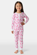 A smiling young child with long dark hair models Roller Rabbit's Kids Gigi Giraffe Pajamas—white Pima cotton pajamas featuring a pink giraffe print—standing barefoot, hand on hip, against a plain white background.