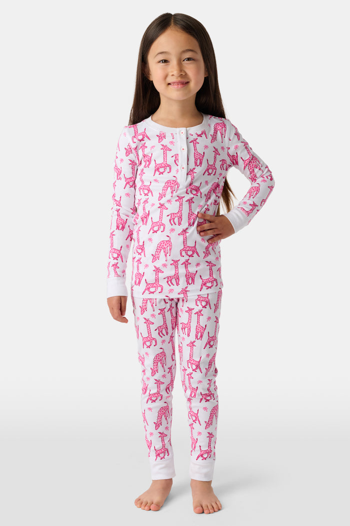 A smiling young child with long dark hair models Roller Rabbit's Kids Gigi Giraffe Pajamas—white Pima cotton pajamas featuring a pink giraffe print—standing barefoot, hand on hip, against a plain white background.