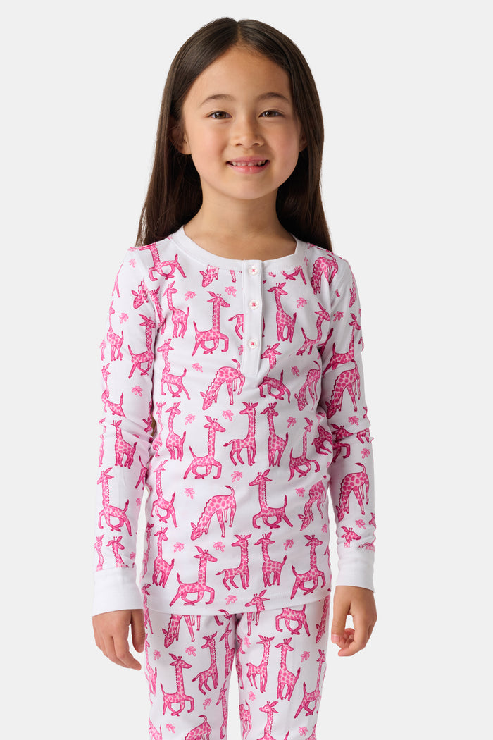A smiling young child with long dark hair models Roller Rabbit's Kids Gigi Giraffe Pajamas—white Pima cotton pajamas featuring a pink giraffe print—standing barefoot, hand on hip, against a plain white background.