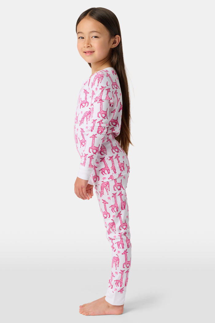 A smiling young child with long dark hair models Roller Rabbit's Kids Gigi Giraffe Pajamas—white Pima cotton pajamas featuring a pink giraffe print—standing barefoot, hand on hip, against a plain white background.