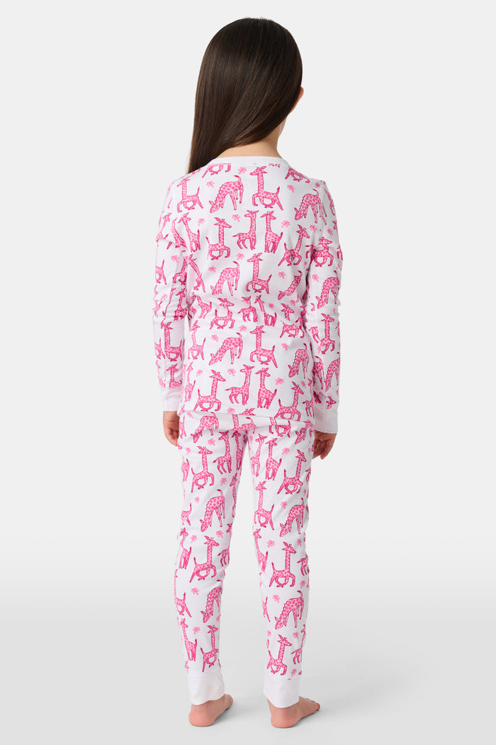 A smiling young child with long dark hair models Roller Rabbit's Kids Gigi Giraffe Pajamas—white Pima cotton pajamas featuring a pink giraffe print—standing barefoot, hand on hip, against a plain white background.