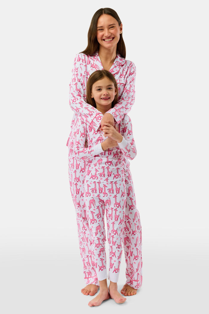 A smiling young child with long dark hair models Roller Rabbit's Kids Gigi Giraffe Pajamas—white Pima cotton pajamas featuring a pink giraffe print—standing barefoot, hand on hip, against a plain white background.