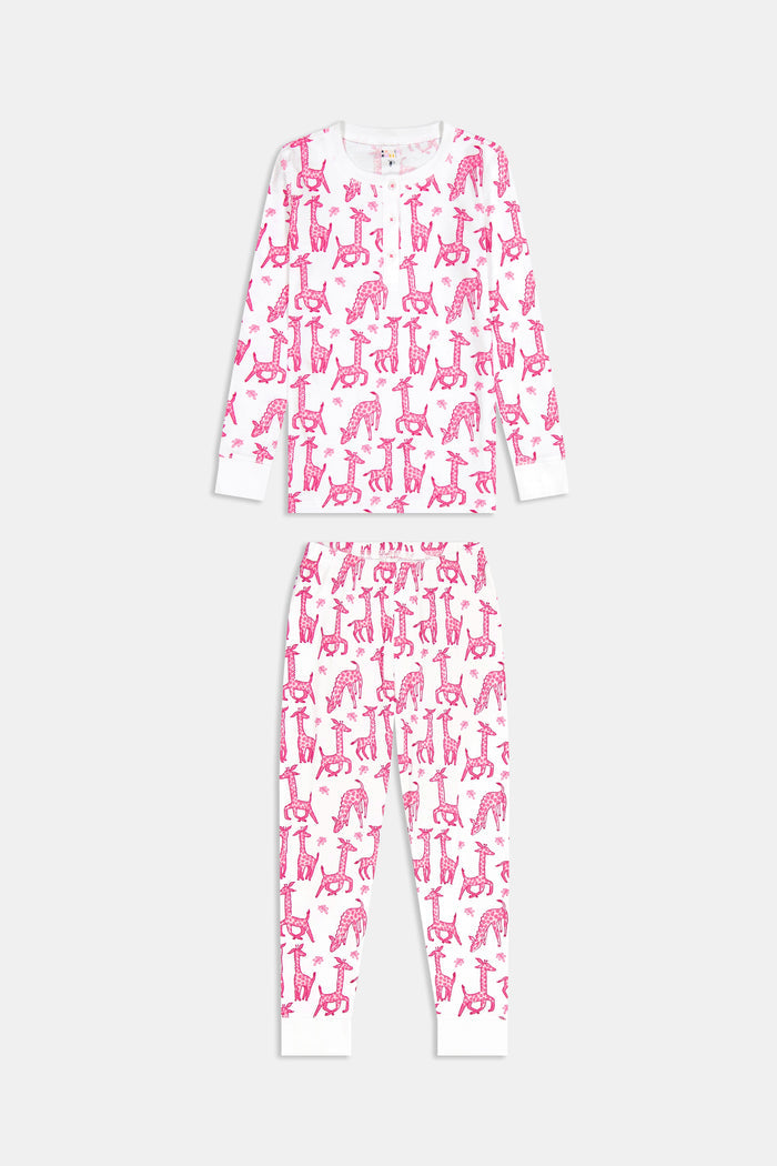 A smiling young child with long dark hair models Roller Rabbit's Kids Gigi Giraffe Pajamas—white Pima cotton pajamas featuring a pink giraffe print—standing barefoot, hand on hip, against a plain white background.