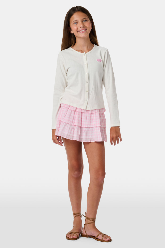 Three girls stand in front of lockers, holding notebooks. Smiling at each other, they are dressed in pink and white outfits, with one wearing the Roller Rabbit Kids Gingham Hearts Sami Skirt. Blue and pink backpacks are visible by the lockers.