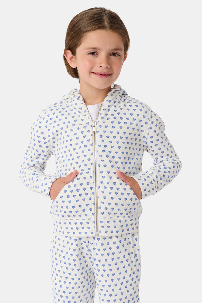 A smiling young child with brown hair wears the Roller Rabbit Kids Hearts Harley Hoodie and matching pants, both made from 100 cotton French terry, standing with hands in the hoodie’s pockets against a plain light background.